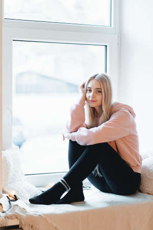 Lovely girl sitting on the windowsill with pillows around thinking and dreaming indoors. She has...