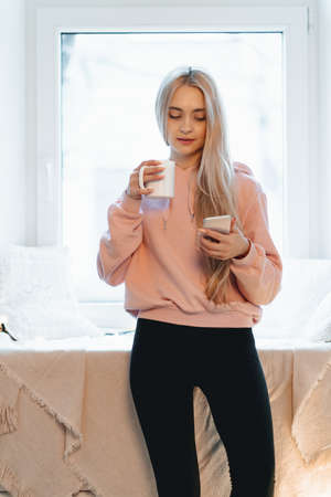 Pretty girl sitting on the window sill with a cup of coffee, tea and smartphone in hands. She has long blonde hair, smile and looking at her phone. Wearing pink, pale parka.の写真素材