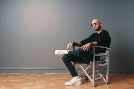 A modern, stylish man sitting leg over leg, in a confident pose on a silver chair in the studio on a gray wall background. He wears a beard, glasses, black casual clothes and white sneakers.の写真素材