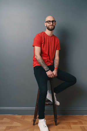 Smiling bearded young man on gray background. Stylish man wearing glasses, looking at camera and smiling while sitting on a tall chair.の写真素材
