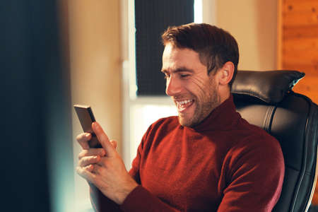 A man happy to receive good news while looking at the smartphone screen, sitting in a leather chair in the officeの写真素材