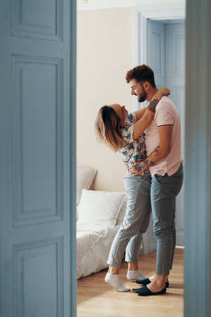 Profile view of loving young couple hugging while standing in their bedroom. View from another room through the doorwayの写真素材