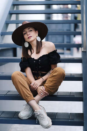 Fashionable brunette woman with long hair, wearing stylish large wicker hat, posing on the stairs. Young beautiful happy smiling fashionable traveller, tourist ladyの写真素材