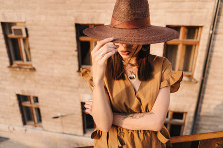 Young fashion girl dressed in a summer dress with a wicker wide hat walks and poses in the old city. Street fashion portrait of young beautiful woman walking in the cityの写真素材