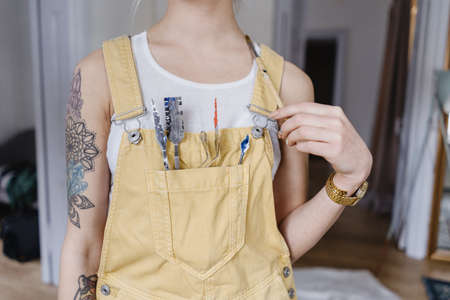 Closeup young female artist holding painting instruments in a pocket on the bust indoors. Tattoos on hands.の写真素材