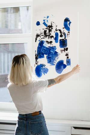 Blonde caucasian woman placing abstract black and phantom blue paint on the wall near the window...