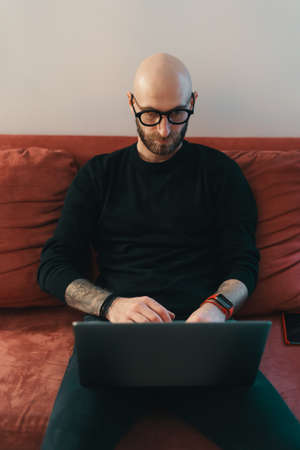 Modern Caucasian man with beard and glasses relaxing while sitting on a couch and surfing internet...