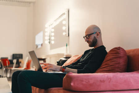Modern caucasian man with beard and glasses relaxing in studio interior, sitting on a coral red...