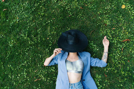 Fashion portrait of a young girl dressed in a blue striped jacket and black hat. Girl lies on a green lawn in a park in the cityの写真素材