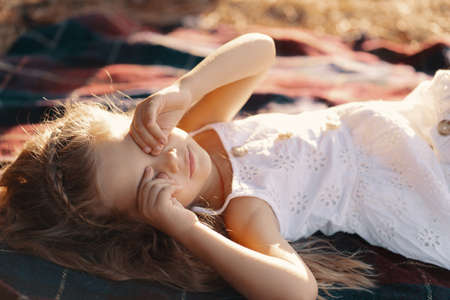 Carefree lovely girl in white dress with blonde wavy hair lying on a blanket enjoying the sun raysの写真素材