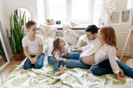Beautiful pregnant woman lying on the bed with her husband and two awesome kids, boy and girl. Bare tummy, jeans, white shirts. Happy momentsの写真素材