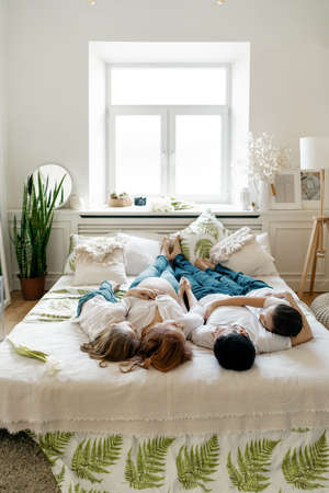 Beautiful pregnant woman lying on the bed with her husband and two awesome kids, boy and girl. Bare tummy, jeans, white shirts. Happy momentsの写真素材