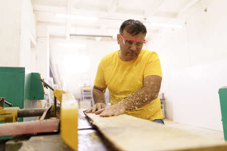 Carpenter cutting a piece of wood for furniture in his woodwork workshop, using a circular saw, and wearing safety goggles.の写真素材