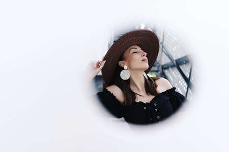 Fashionable brunette woman with long hair, wearing stylish large wicker hat, posing against a white wall with holes. Young beautiful happy smiling fashionable traveller, tourist ladyの写真素材