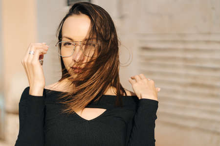 Attractive young brunette woman wearing a black top and transparent glasses posing on the street....