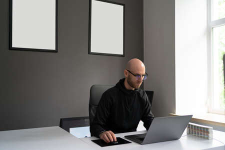Young man with glasses working concentrated at laptop in his modern office with copy space in frames on the wallの写真素材