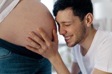 A happy smiling man is watching his beautiful pregnant wifes bare tummy touching it tenderlyの写真素材
