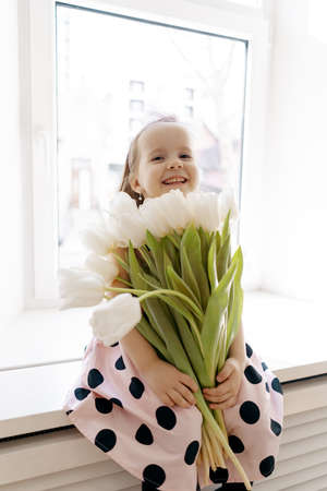 Close up charming little girl smiling, holding white tulips near the window, day light reflecting on her beautiful faceの写真素材