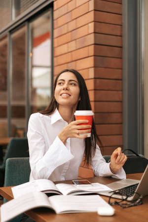 Smiling businesswoman enjoying take away coffee while working in a cafe on a portable computer...