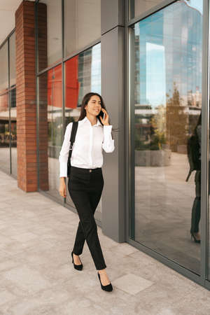 Woman with business backpack walking while speaking on the phone near a business buildingの写真素材