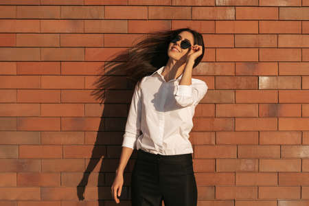 Pretty brunette woman portrait on brick background with shadow in white shirt and sun glasses...