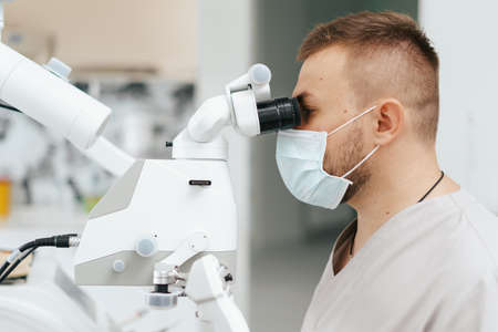 Young man with patient bib on a dental chair and a dentist who sits next to him. He looks on his teeth using a dental microscope and holds a dental bur and a mirror.の写真素材