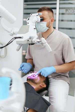 Young man with patient bib on a dental chair and a dentist who sits next to him. He looks on his teeth using a dental microscope and holds a dental bur and a mirror.の写真素材