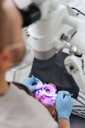 Young man with patient bib on a dental chair and a dentist who sits next to him. He looks on his teeth using a dental microscope and holds a dental bur and a mirror.の写真素材