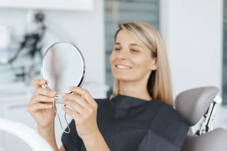 Young woman patient is looking in the mirror and admires her new smile after dental treatment in the dentistry clinic.の写真素材