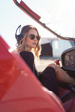 Young fashionable woman pilot in headset ready to fly in small red airplane. Beautiful life, aristocratic lady in black dress in blonde wavy hair.の写真素材