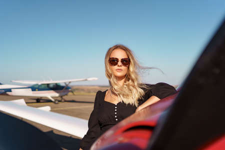 Beautiful blonde woman wearing modern sunglasses posing near a red private plane with sky in...
