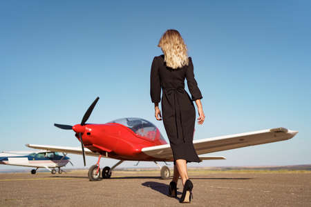 Back view Fashionable women in beautiful classy black dress walking to a red private plane and blue sky in backgroundの写真素材