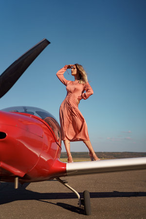 Pretty blond woman with wavy hair wearing pink silk classy dress posing near a private planeの写真素材