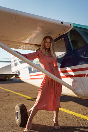 Pretty blond woman with wavy hair wearing pink silk classy dress posing near a private planeの写真素材