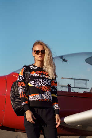 Young woman model with a modern haircut and fashionable sunglasses posing near a red plane wearing...