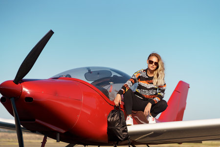 Young woman model with a modern haircut and fashionable sunglasses posing near a red plane wearing...