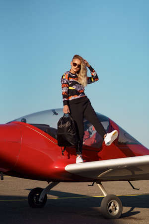 Carefree woman model with a modern haircut and fashionable sunglasses posing near a red plane...