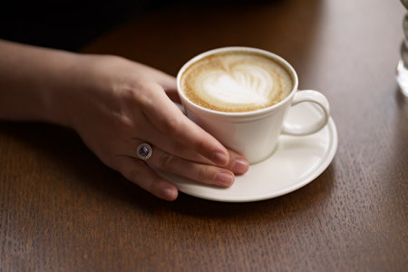 Female hands holding a white cup of delicious coffee with foam over wooden table, side view....