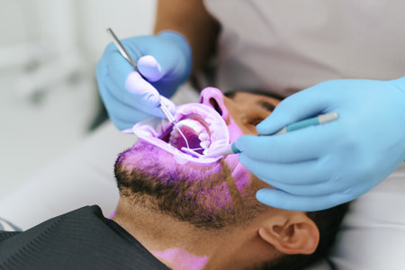 Young man with patient bib on a dental chair, pink light oriented on his teeth and a dentist who sits next to him wearing blue medical gloves while examining oral cavityの写真素材