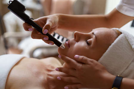 View of doctor cosmetologist doing anti aging procedure in cosmetology office. Satisfied woman in disposable hat lying on couch and relaxing. Working with Apparatus.の写真素材