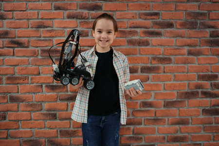 Smiling School boy holding a robot and its remote control on brick wall background