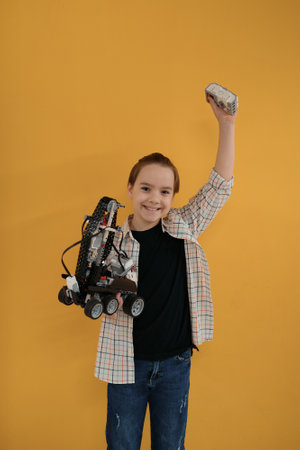 Smiling School boy holding a robot and its remote control on yellow wall background