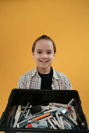 Smiling School boy holding a box with plastic robotic peaces on yellow wall background. Ready to...