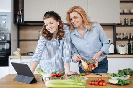 Mom and daughter cooking together in the kitchen using a receipt on tablet. They use vegetables for cooking in a good mood, happy to be togetherの写真素材