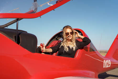 Young fashionable woman pilot in headset ready to fly in small red airplane. Beautiful life, aristocratic lady in black dress in blonde wavy hair.の写真素材