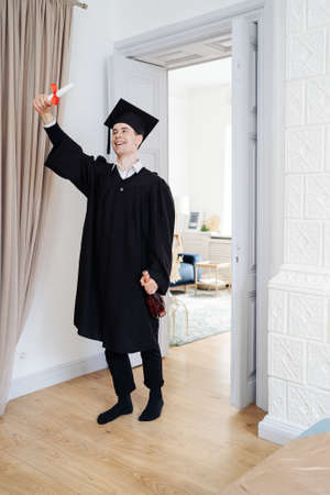 Education, graduation and people concept - happy male student with diploma at home showing his...