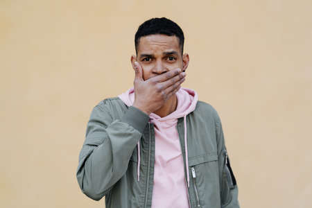 Young handsome Afro man on yellow background cover mouth with hands, expression of surprise, secret conceptの写真素材