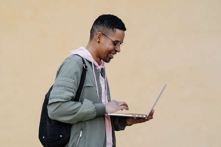 African man in glasses holding laptop on pale yellow background. Shot of carefree black male freelancer smiling to camera.の写真素材