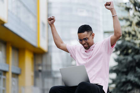 Handsome young man in glasses celebrate victory with laptop outdoors on a background of corporate buildingの写真素材