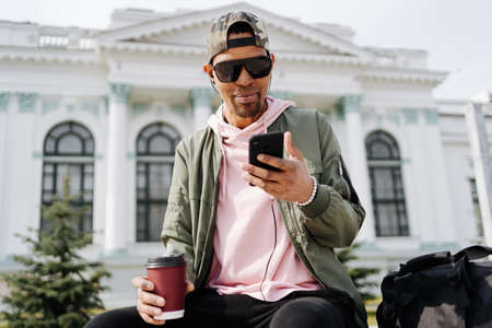 People, technology, travel and tourism - Afro man using smartphone on city street bench and texting while drinking coffeeの写真素材